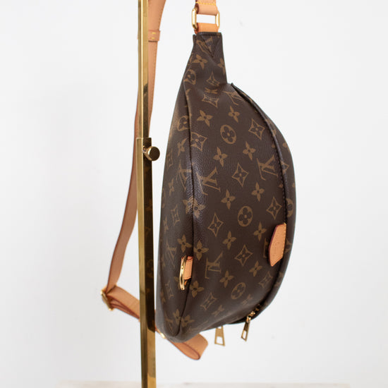 A Louis Vuitton monogram canvas sling bag with tan leather trim, hanging from a gold metal display stand against a white studio background.
