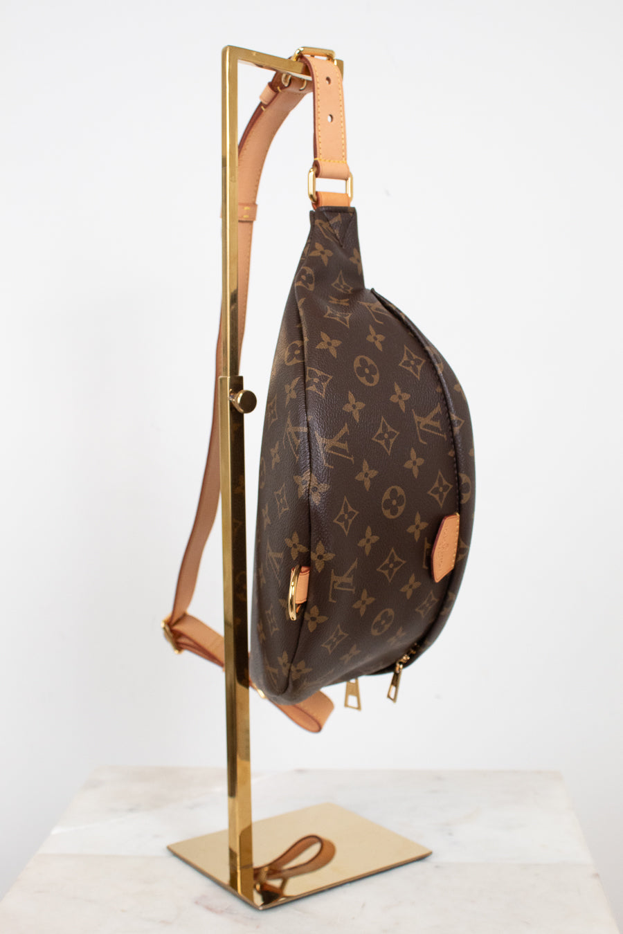 A Louis Vuitton monogram canvas sling bag with tan leather trim, hanging from a gold metal display stand against a white studio background.