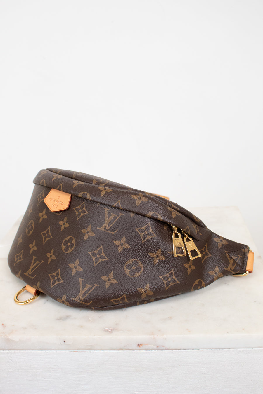 A Louis Vuitton monogram canvas fanny pack with gold-tone hardware, resting on a white marble surface against a white background.
