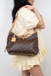 A woman wearing a yellow and white gingham dress carries a Louis Vuitton brown monogram shoulder bag with tan leather trim and strap.
