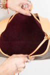 Close-up of a handbag interior with a tan leather rim and a dark burgundy velvet lining, held open by two hands wearing rings and bracelets.