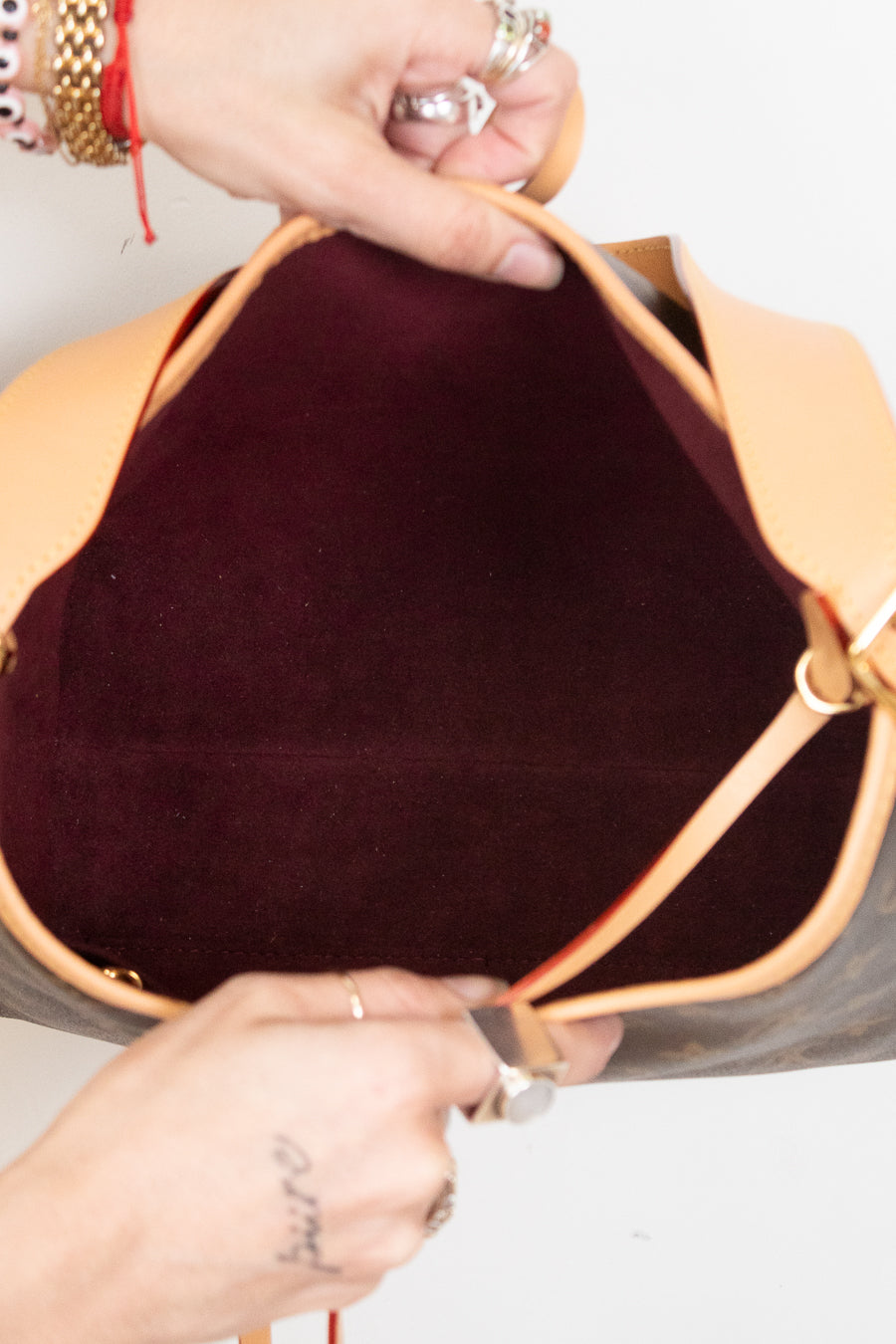 Close-up of a handbag interior with a tan leather rim and a dark burgundy velvet lining, held open by two hands wearing rings and bracelets.