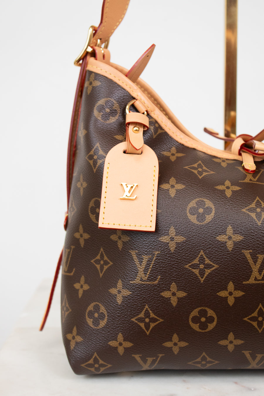 A close-up of a Louis Vuitton handbag in brown monogram canvas with light tan leather trim and a matching luggage tag featuring a gold-tone LV logo.