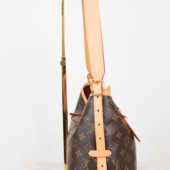 A Louis Vuitton monogram bucket bag in brown coated canvas with tan leather trim and strap, displayed upright on a gold stand against a white background.