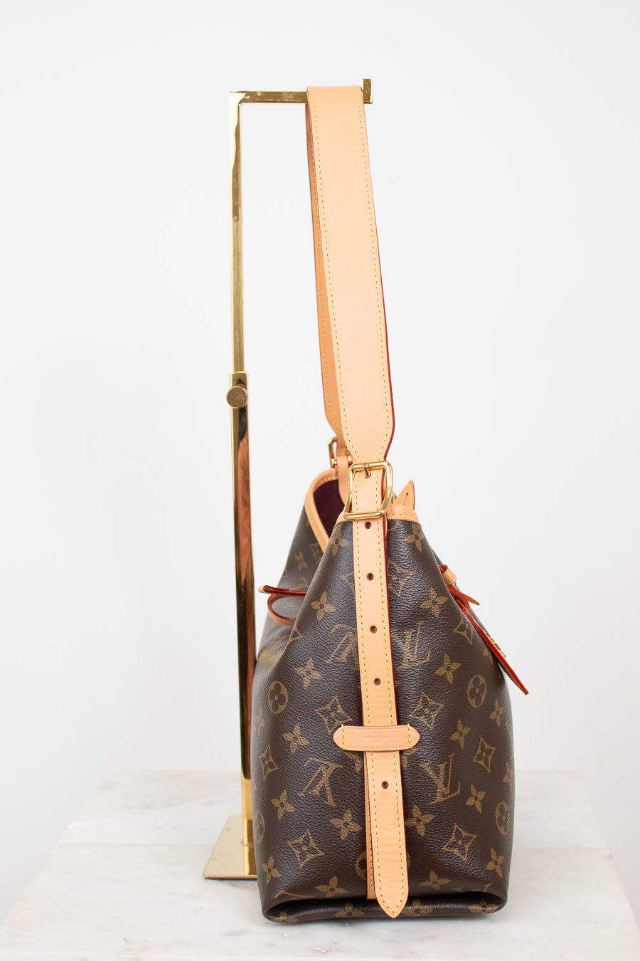 A Louis Vuitton monogram bucket bag in brown coated canvas with tan leather trim and strap, displayed upright on a gold stand against a white background.