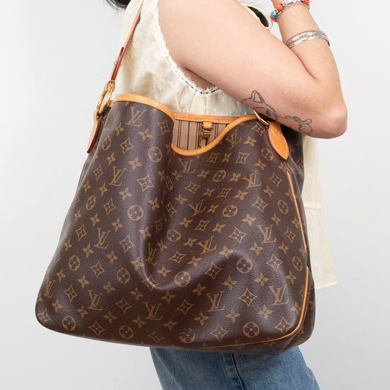 A woman wearing a cream lace-trim top and blue jeans carries a brown Louis Vuitton monogram shoulder bag with tan leather trim against a light gray background.