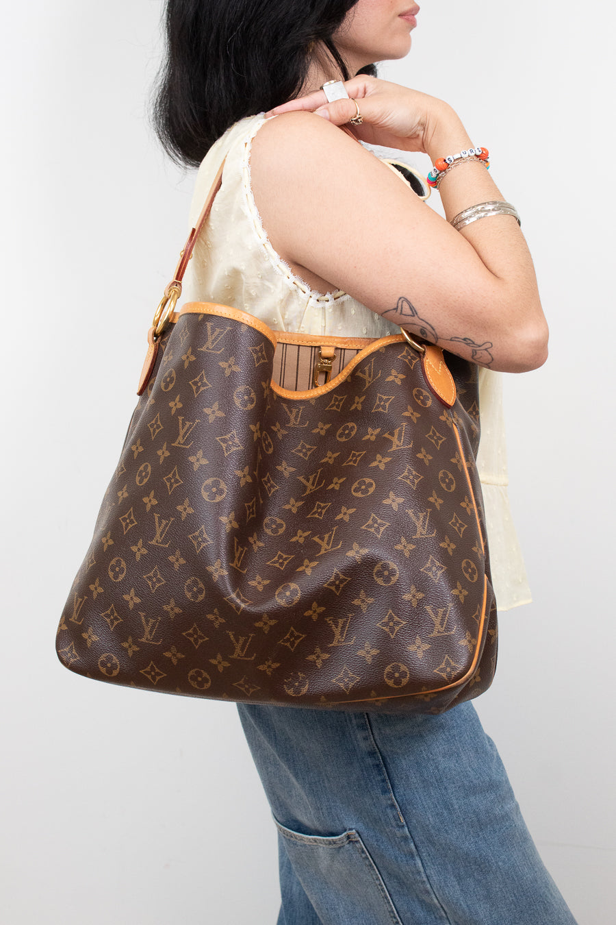 A woman wearing a cream lace-trim top and blue jeans carries a brown Louis Vuitton monogram shoulder bag with tan leather trim against a light gray background.