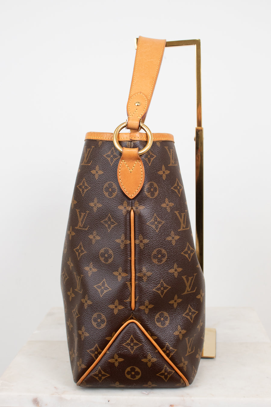 Louis Vuitton monogram coated canvas shoulder bag with tan leather trim and a gold-tone metal ring detail, displayed on a white marble surface with a gold stand against a white background.
