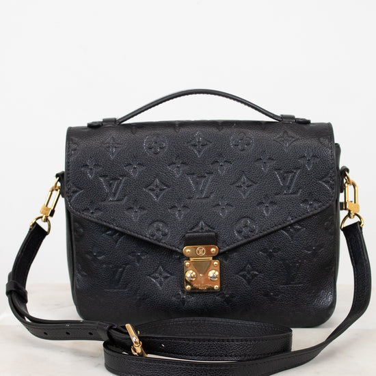 A black embossed monogram shoulder bag with gold-tone hardware, featuring a top handle, a detachable shoulder strap, and a front flap secured by a gold clasp, displayed on a light marble surface against a white background.