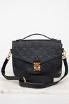 A black embossed monogram shoulder bag with gold-tone hardware, featuring a top handle, a detachable shoulder strap, and a front flap secured by a gold clasp, displayed on a light marble surface against a white background.