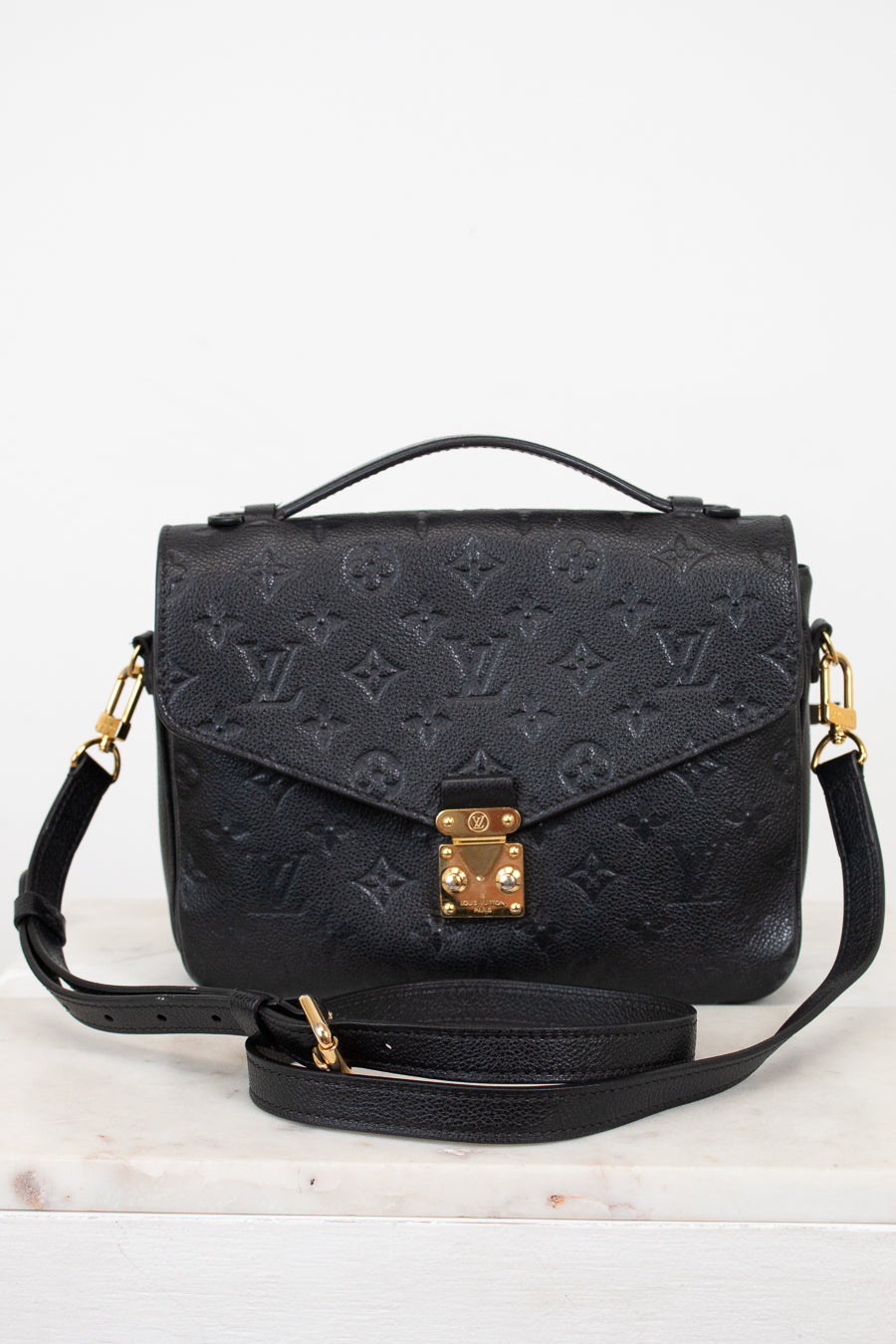 A black embossed monogram shoulder bag with gold-tone hardware, featuring a top handle, a detachable shoulder strap, and a front flap secured by a gold clasp, displayed on a light marble surface against a white background.