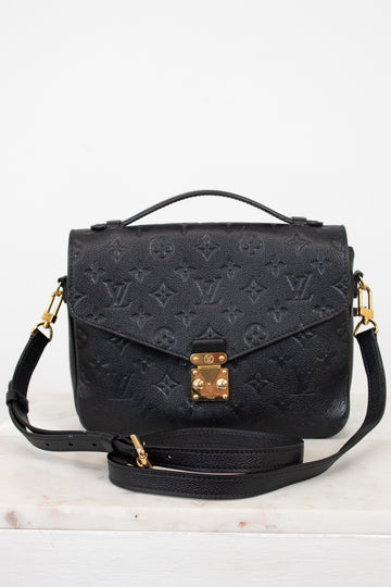 A black embossed monogram shoulder bag with gold-tone hardware, featuring a top handle, a detachable shoulder strap, and a front flap secured by a gold clasp, displayed on a light marble surface against a white background.