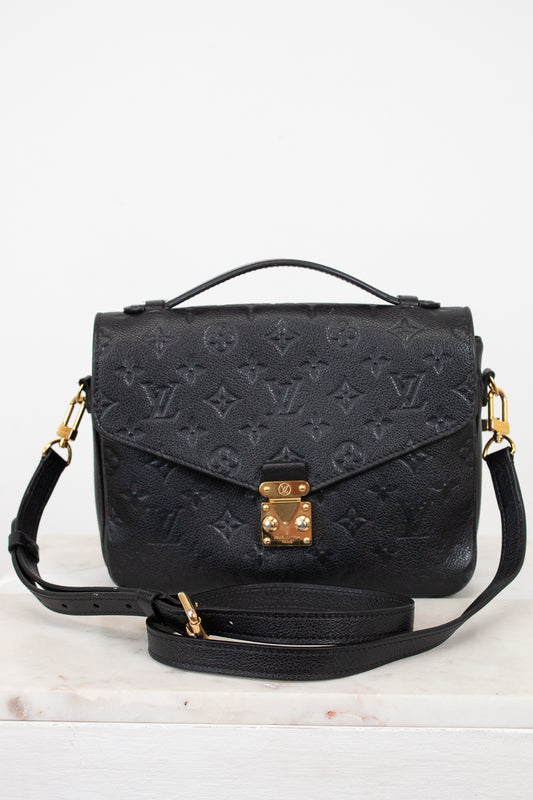 A black embossed monogram shoulder bag with gold-tone hardware, featuring a top handle, a detachable shoulder strap, and a front flap secured by a gold clasp, displayed on a light marble surface against a white background.