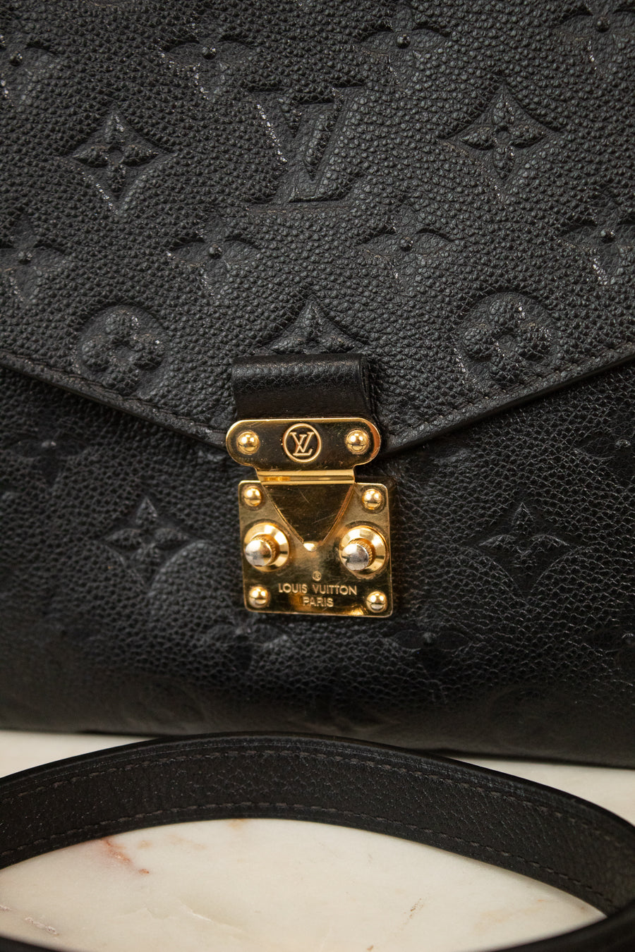 Close-up of a black Louis Vuitton monogram bag with a gold-tone closure  lock and strap on a light marble surface.