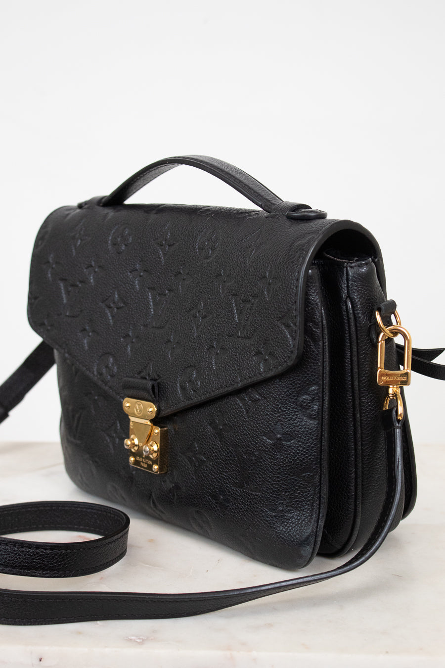A black embossed leather handbag with a top handle, gold-tone clasp, and matching shoulder strap, photographed on a light neutral background.