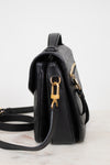 A black textured leather shoulder bag shown from a side profile view, featuring gold-tone hardware and a matching black strap on a light marble surface against a white background.
