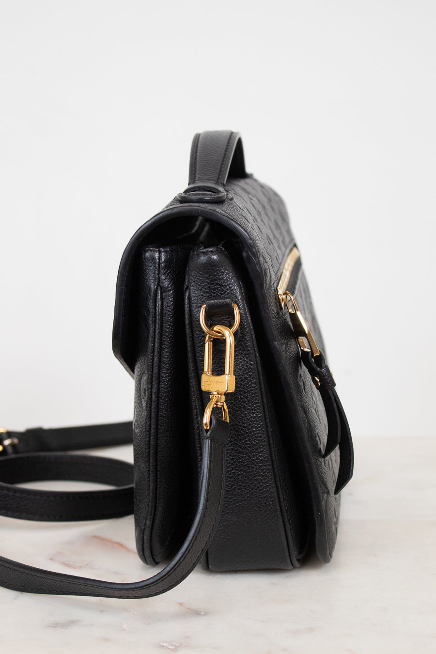 A black textured leather shoulder bag shown from a side profile view, featuring gold-tone hardware and a matching black strap on a light marble surface against a white background.
