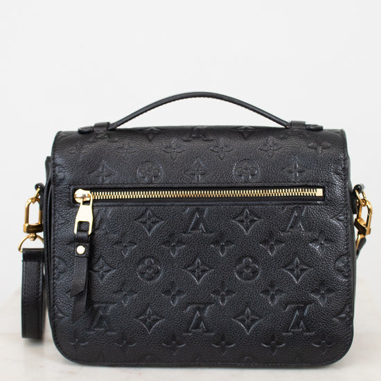 A black embossed monogram shoulder bag with gold-tone hardware, featuring a top handle, a back zip pocket, and a detachable shoulder strap, displayed on a light marble surface against a white background.