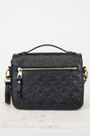 A black embossed monogram shoulder bag with gold-tone hardware, featuring a top handle, a back zip pocket, and a detachable shoulder strap, displayed on a light marble surface against a white background.