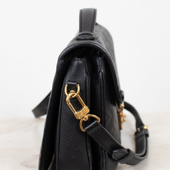 A black pebbled leather crossbody bag with gold-tone hardware, shown from a side profile view on a light marble surface against a white background.