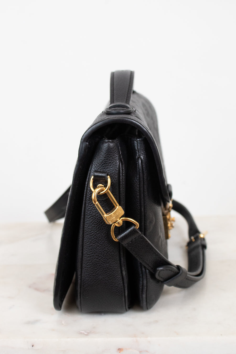 A black pebbled leather crossbody bag with gold-tone hardware, shown from a side profile view on a light marble surface against a white background.