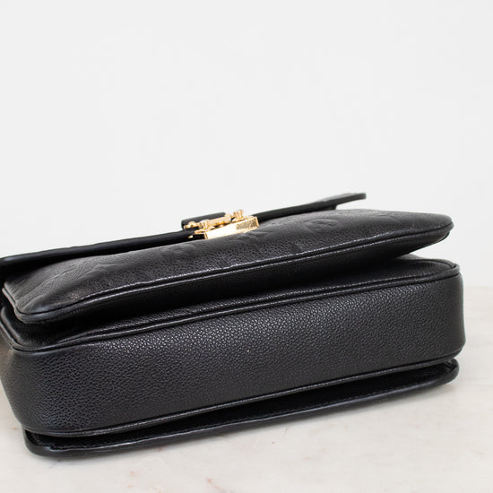 Bottom view of a black pebbled leather bag with a gold-tone metal clasp, resting on a light marble surface against a white background.