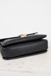 Bottom view of a black pebbled leather bag with a gold-tone metal clasp, resting on a light marble surface against a white background.