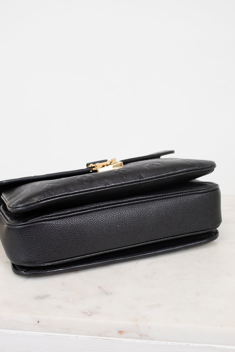 Bottom view of a black pebbled leather bag with a gold-tone metal clasp, resting on a light marble surface against a white background.