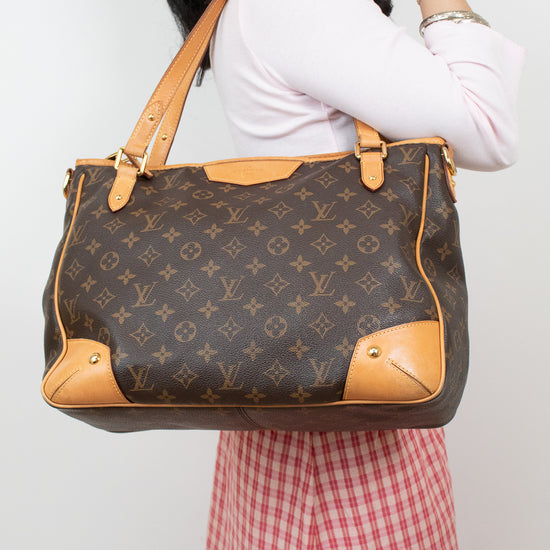 A woman wearing a white long-sleeve top and a red-and-white gingham skirt carries a large Louis Vuitton monogram tote bag with tan leather trim and shoulder straps against a plain light background.