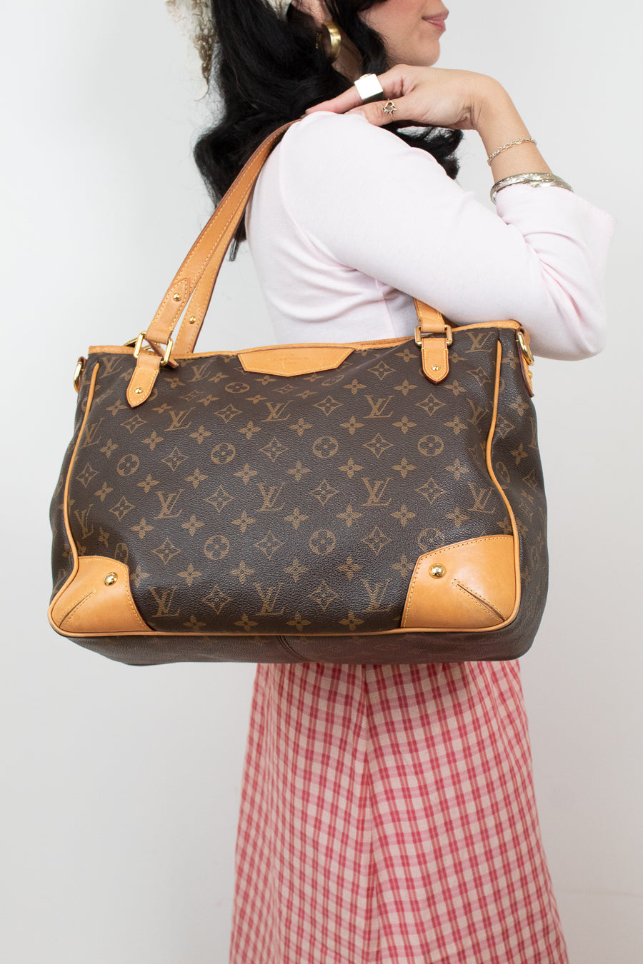 A woman wearing a white long-sleeve top and a red-and-white gingham skirt carries a large Louis Vuitton monogram tote bag with tan leather trim and shoulder straps against a plain light background.