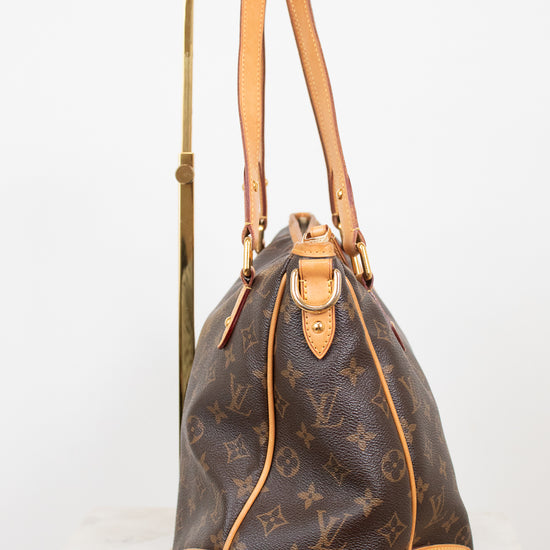 Side view of a Louis Vuitton monogram coated canvas tote bag with tan leather trim and handles, displayed on a gold metal stand against a white background.