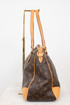 Side view of a Louis Vuitton monogram coated canvas tote bag with tan leather trim and handles, displayed on a gold metal stand against a white background.