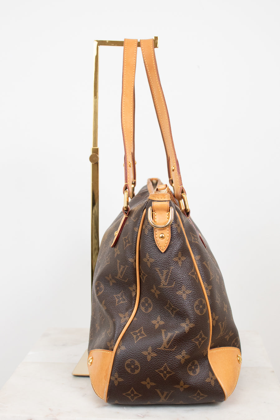 Side view of a Louis Vuitton monogram coated canvas tote bag with tan leather trim and handles, displayed on a gold metal stand against a white background.
