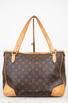 Louis Vuitton monogram canvas tote bag with tan leather handles and corner protectors, displayed on a stand against a white background.