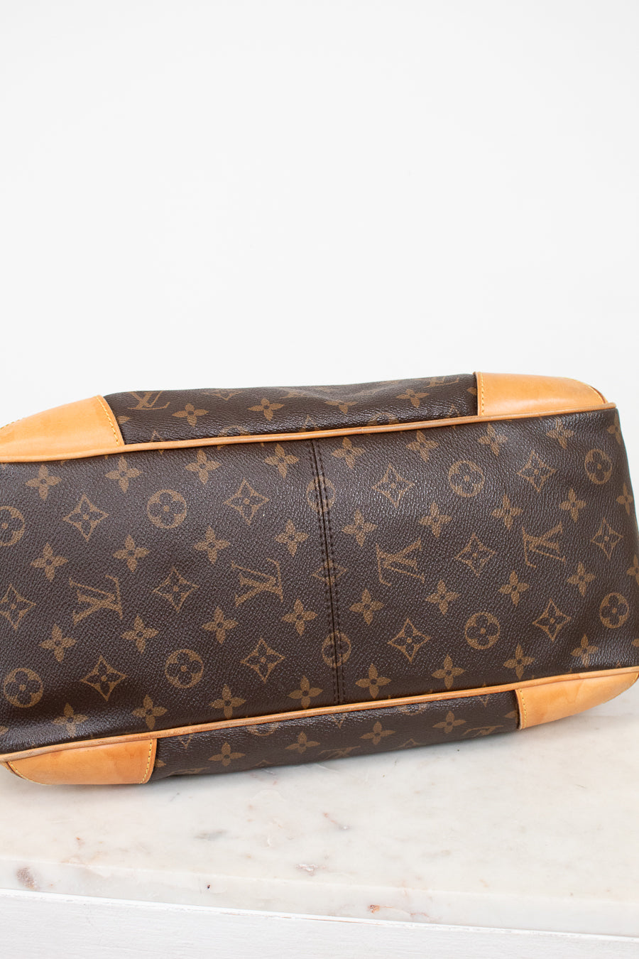 Louis Vuitton monogram coated canvas handbag shown from the bottom corner, featuring brown coated canvas with tan leather trim and corner protectors on a light marble surface.