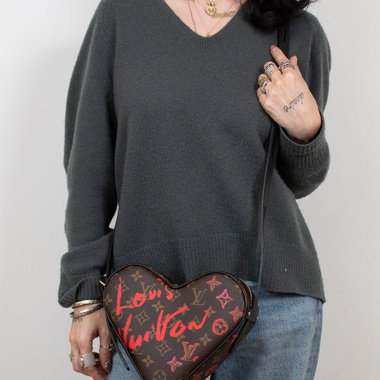 Person wearing a dark gray sweater and blue jeans, holding a heart-shaped Louis Vuitton bag.