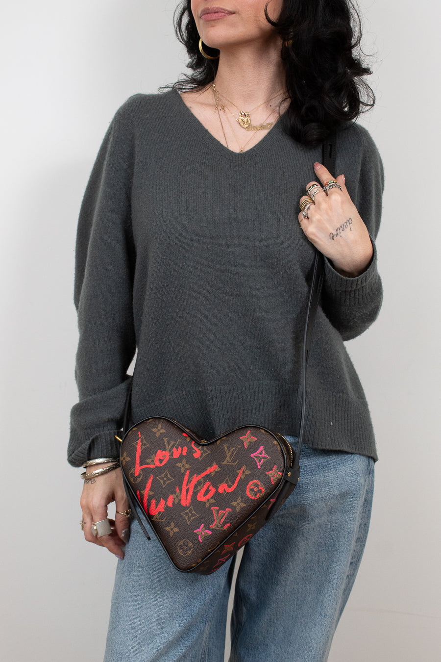 Person wearing a dark gray sweater and blue jeans, holding a heart-shaped Louis Vuitton bag.