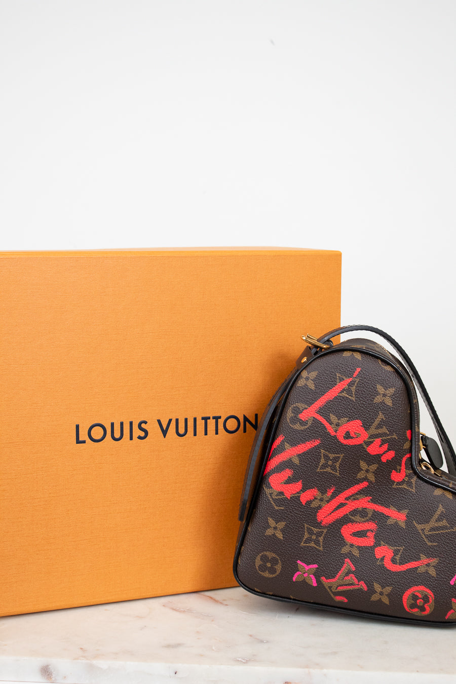 Louis Vuitton handbag with red and gold monogram pattern next to an orange box on a white background