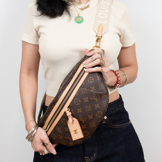 A woman wearing a cream short-sleeve top and a dark denim mini skirt carries a Louis Vuitton monogrammed crescent-shaped crossbody/bum bag. The bag features a brown monogram pattern with tan leather trim and a cream canvas strap printed with the name 'LOUIS VUITTON'.