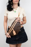 A woman wearing a cream short-sleeve top and a dark denim mini skirt carries a Louis Vuitton monogrammed crescent-shaped crossbody/bum bag. The bag features a brown monogram pattern with tan leather trim and a cream canvas strap printed with the name 'LOUIS VUITTON'.