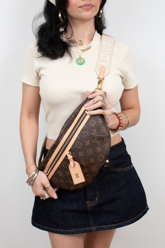 A woman wearing a cream short-sleeve top and a dark denim mini skirt carries a Louis Vuitton monogrammed crescent-shaped crossbody/bum bag. The bag features a brown monogram pattern with tan leather trim and a cream canvas strap printed with the name 'LOUIS VUITTON'.