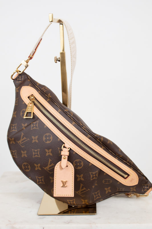 Louis Vuitton monogram coated canvas fanny pack with tan leather trim and gold-tone hardware, displayed on a gold stand against a white background.