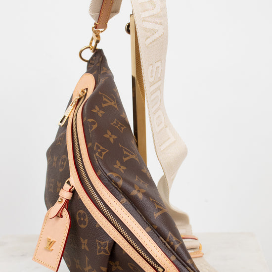 Louis Vuitton monogram coated canvas sling bag with beige leather trim and a wide cream canvas shoulder strap, displayed on a gold stand against a white background.