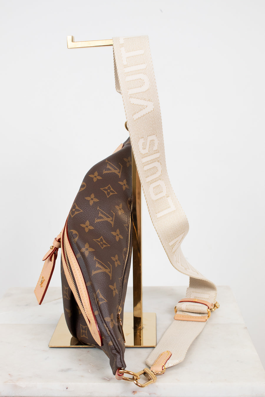 Louis Vuitton monogram shoulder bag in brown coated canvas with a beige canvas strap featuring white 'LOUIS VUITTON' text, displayed on a gold stand against a white background.