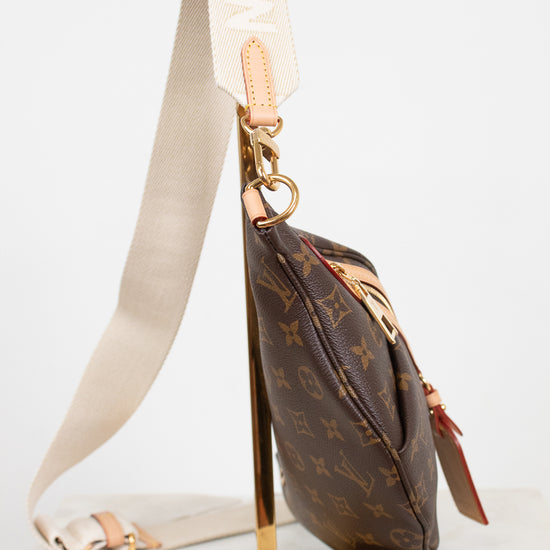 A Louis Vuitton monogram shoulder bag in brown coated canvas with gold-tone hardware, displayed on a gold metal stand. The bag features a cream-colored fabric strap with white lettering and a small leather patch.