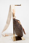 A Louis Vuitton monogram shoulder bag in brown coated canvas with gold-tone hardware, displayed on a gold metal stand. The bag features a cream-colored fabric strap with white lettering and a small leather patch.