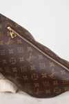 A close-up of a Louis Vuitton monogram coated canvas bag in dark brown with gold-tone zippers and hardware, resting on a light marble surface against a white background.