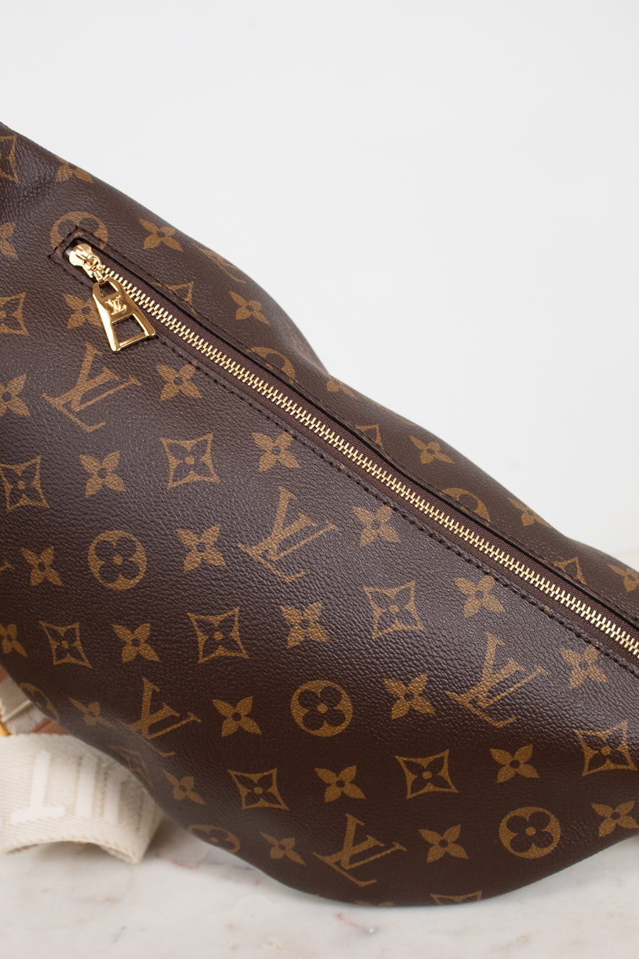 A close-up of a Louis Vuitton monogram coated canvas bag in dark brown with gold-tone zippers and hardware, resting on a light marble surface against a white background.