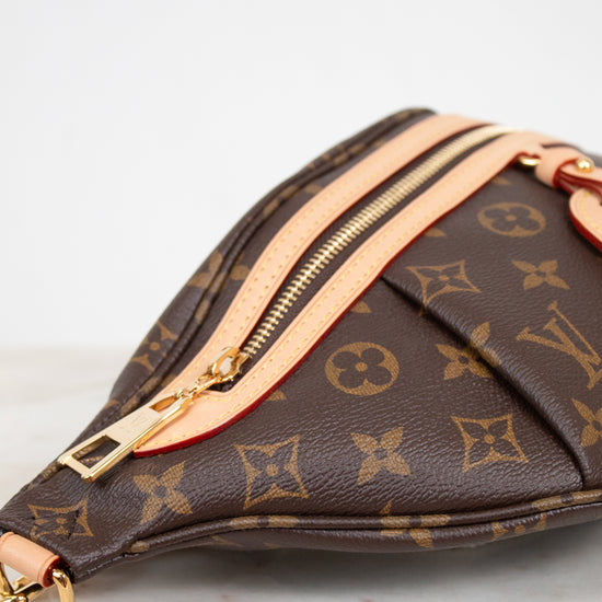 A close-up of a Louis Vuitton monogram bag in brown coated canvas with gold-tone hardware and light beige leather trim, resting on a light marble surface against a white background.