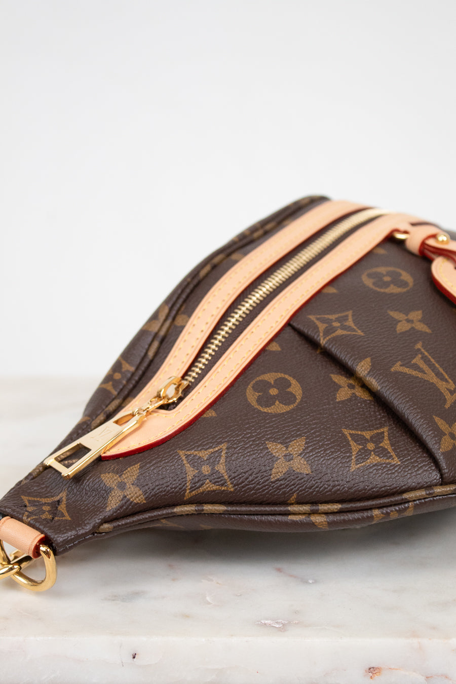 A close-up of a Louis Vuitton monogram bag in brown coated canvas with gold-tone hardware and light beige leather trim, resting on a light marble surface against a white background.